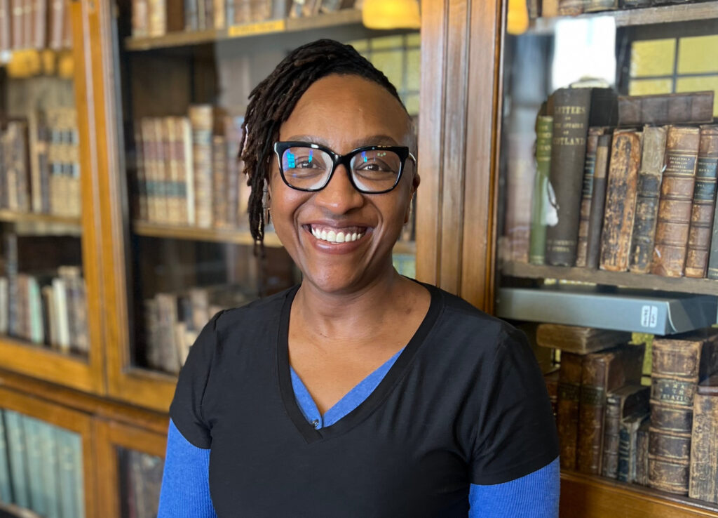 A photo of Liz Mburu standing in front of a wood bookshelf filled with old books.