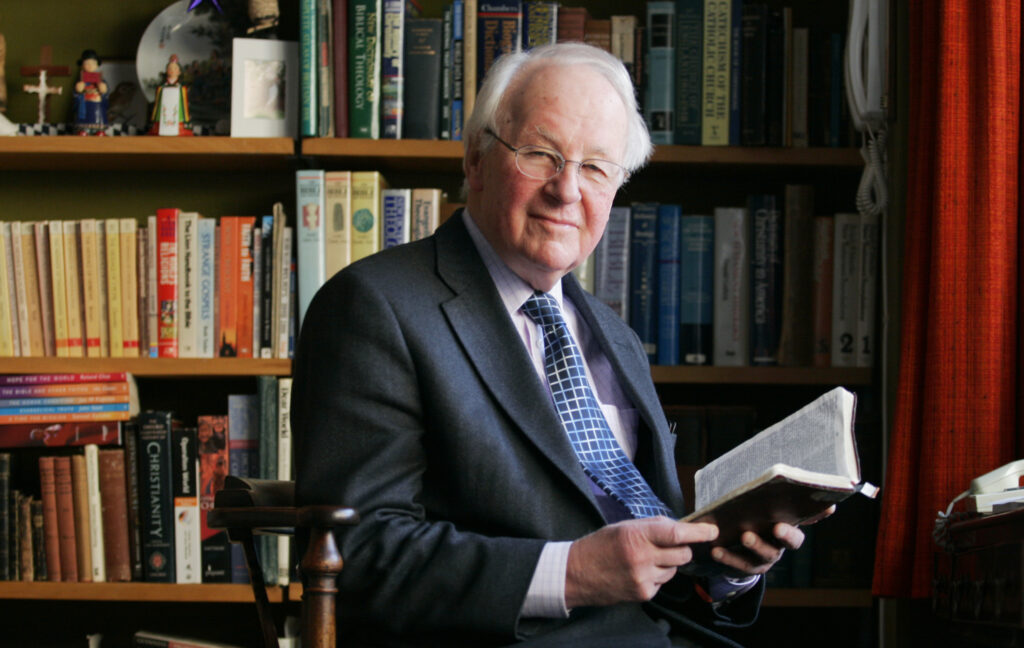 John Stott standing in front of a packed bookshelf with a book in his hand at his All Soul's home. 